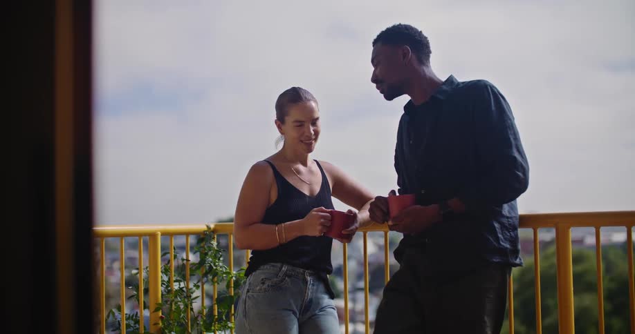 A man and woman stand on a balcony leaning on a railing with mugs in their hands as they talk and the man kisses the woman
