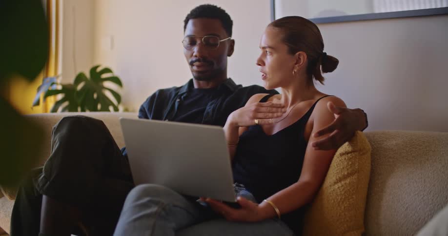 A woman is sitting on the couch with a laptop computer and talking to the man sitting next to her with his arm around her shoulder
