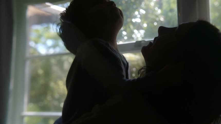 Mother holding toddler son is silhouetted against the windows