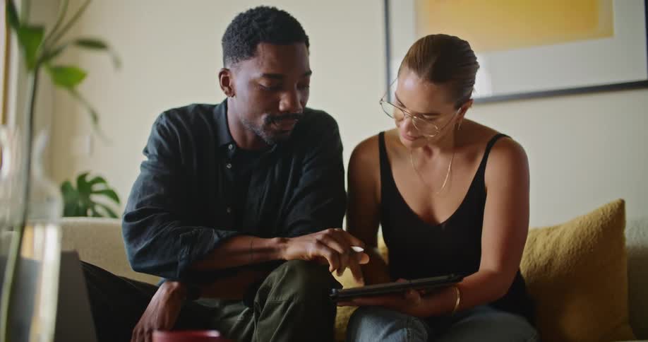 Man and woman sit next to each other on a couch the woman with a tablet on her lap that they are both looking at as the man writes on it with a stylus