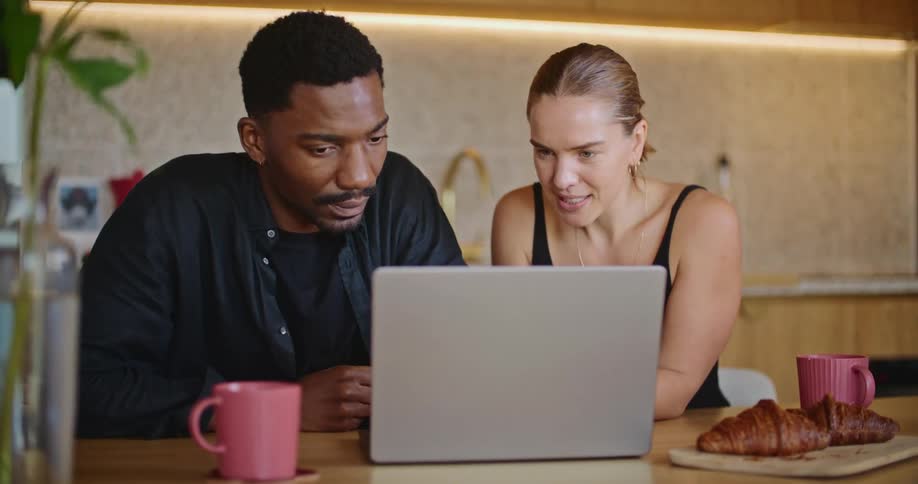 A man an woman sit together in a kitchen while reviewing information on a laptop screen