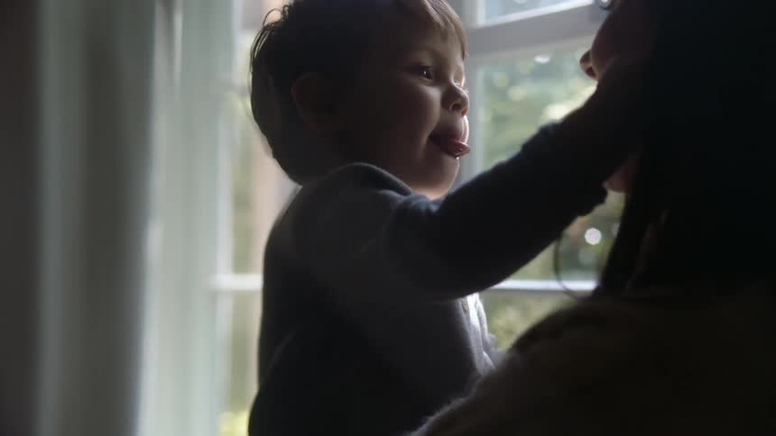 Mother and son sit together in front of a window while the boy tries to lick to the mothers face