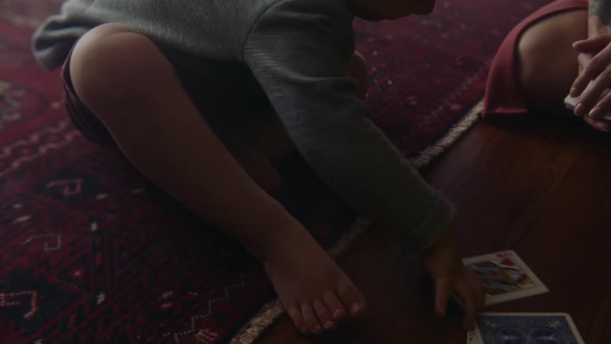 A little boy is sitting on a rug on the floor with another person playing with cards as he has his hand on the queen of hearts