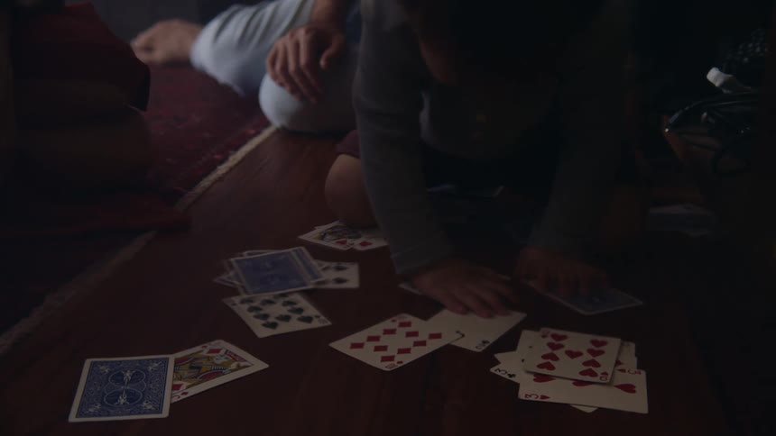 A little boy sits on the ground playing with cards with his father and mother in the background