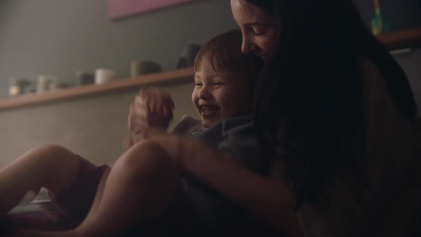 A woman playfully moves the hands of a laughing boy sitting in her lap while sitting indoors during the day
