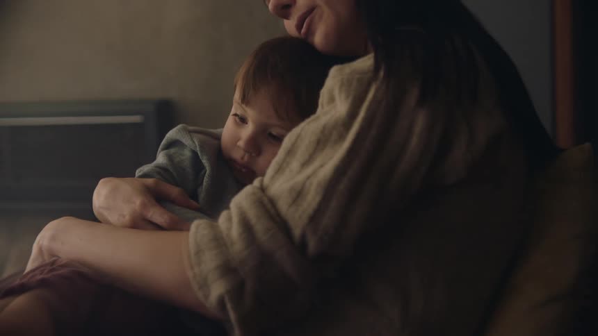 A young woman with long dark hair rocks her young son while he sits on her lap