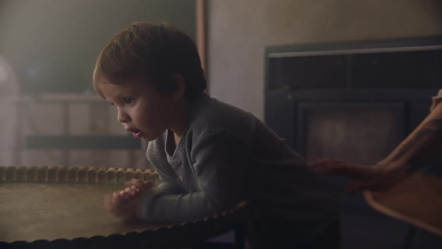 A toddler boy sits and leans over a table while his mother scratches his back