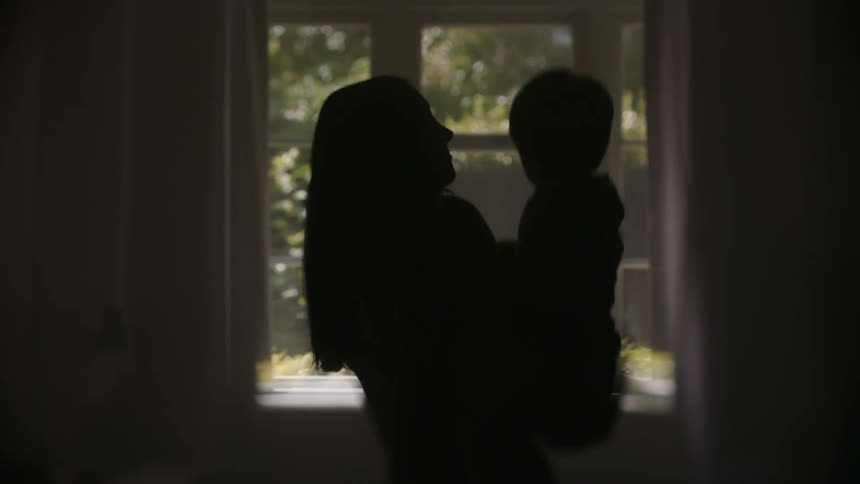 A woman is standing near the window holding her young son as they look at the trees outside