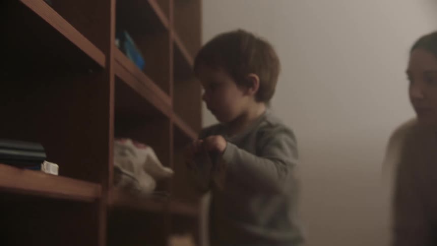 A little boy is picking up playing cards from the floor and putting them on a shelf and his mother is kneeling on the floor helping him