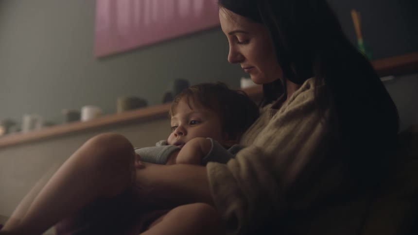 A woman sitting indoors kisses the head of a young boy while rocking him in her lap