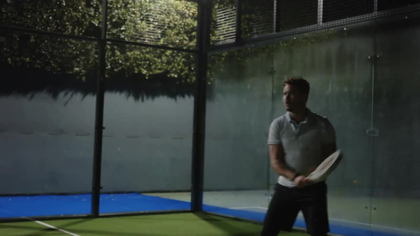 A man plays tennis on a court at night by using a racket to hit a ball