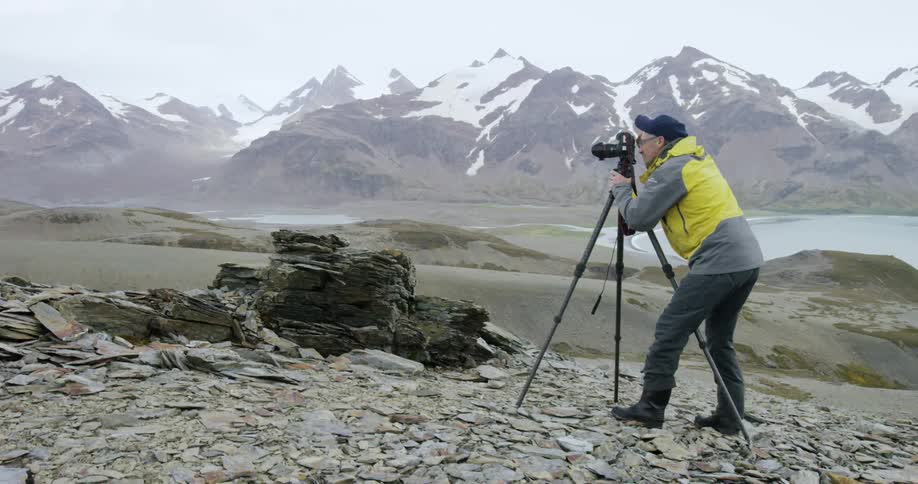 A photographer taking pictures from the top of a rocky hill near snow-capped mountains