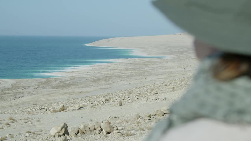 A woman wearing a hat and scarf looks out over a rocky beach during the day