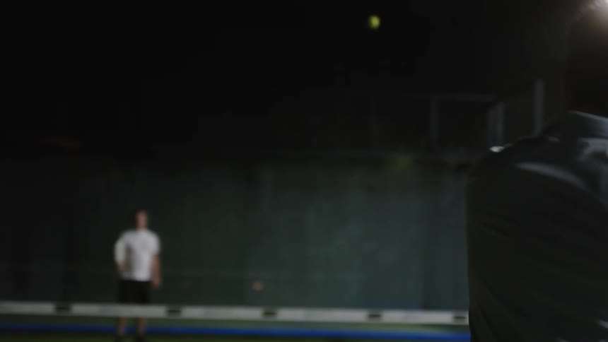 Man with light blue polo shirt plays tennis on tennis court at night