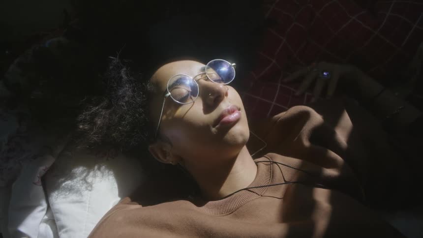 Close up of the face of a young woman wearing glasses resting on her bed under sunlight
