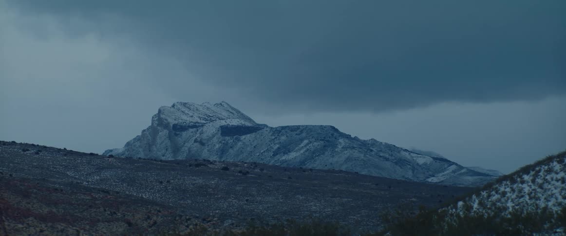 Fresh snow covers a distant desert mountain under dark clouds at dusk