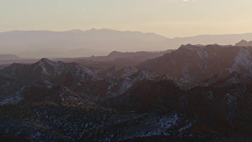An aerial of a large red rock mountain range with peaks and sun shining over the landscape