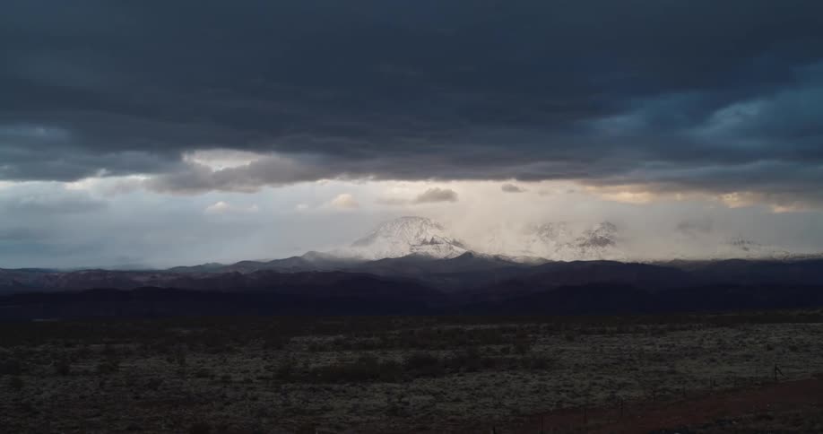 Dense dark clouds deposit snow over a large mountain within the desert in the setting sun