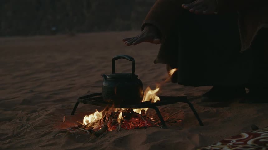 Hand of a person held over the fire under a teapot in the desert sands at evening