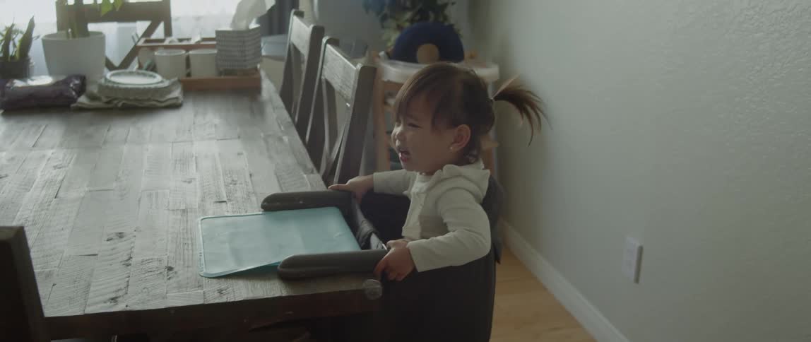 Toddler girl sitting in highchair at dining table while her parents walk by