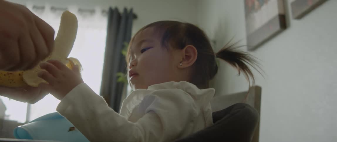 An adult peels a banana and gives it to a baby girl who begins to eat it