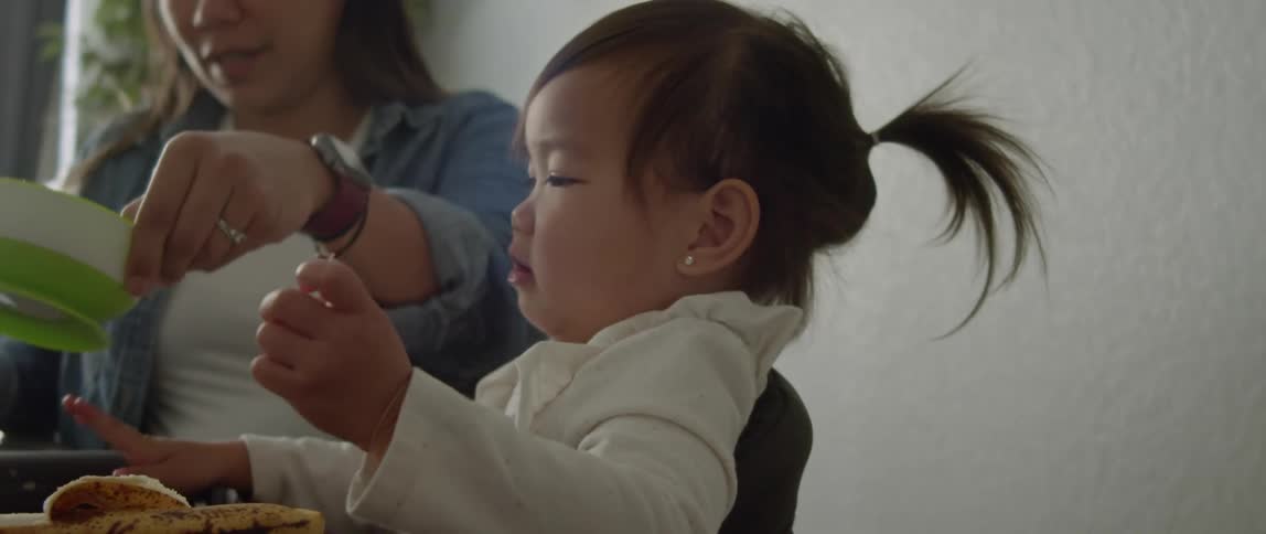 Pregnant mother with denim jacket and white top feeds toddler daughter sitting at table