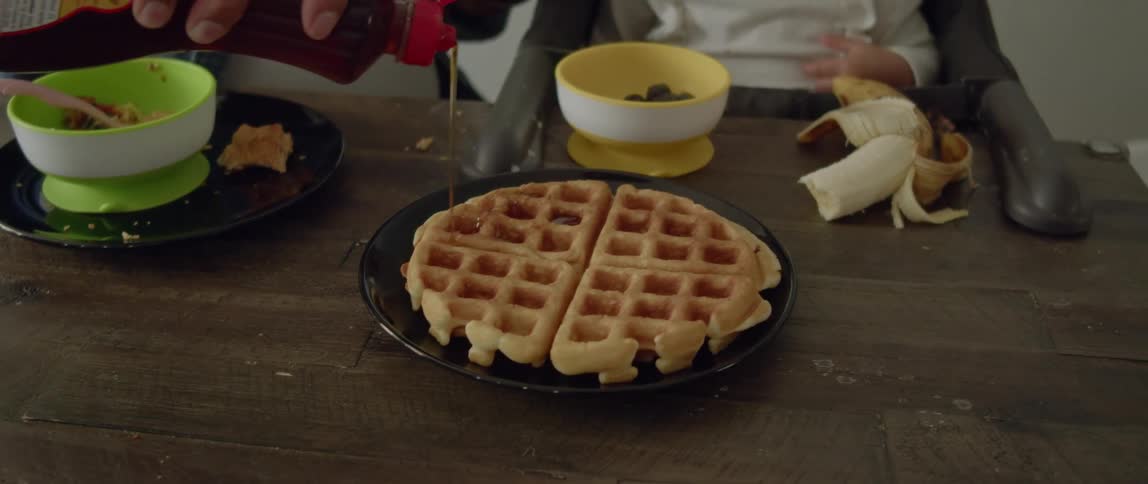 A young mother pours maple syrup over a belgian waffle for her young daughter at breakfast