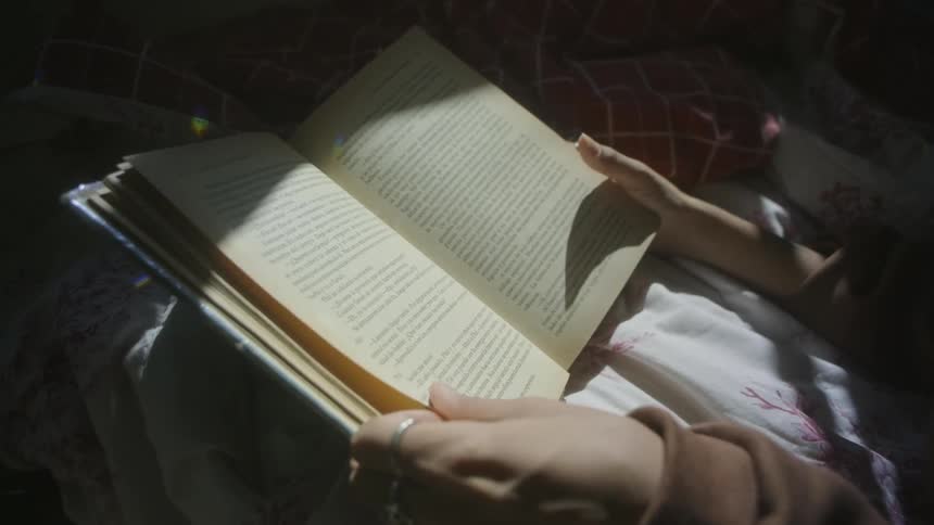 A woman sitting down and reading a book in her lap uses her hands to turn the page