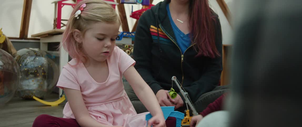 Adults watch a pair of small children play with toys as they all sit together on the floor indoors
