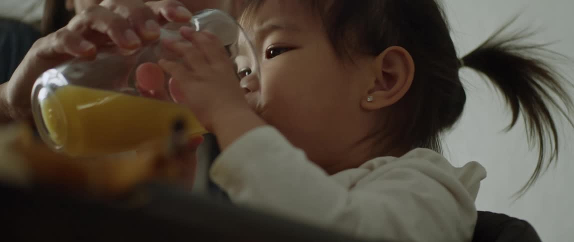 Close up of a mother helping her daughter drink orange juice from a glass in the dining table