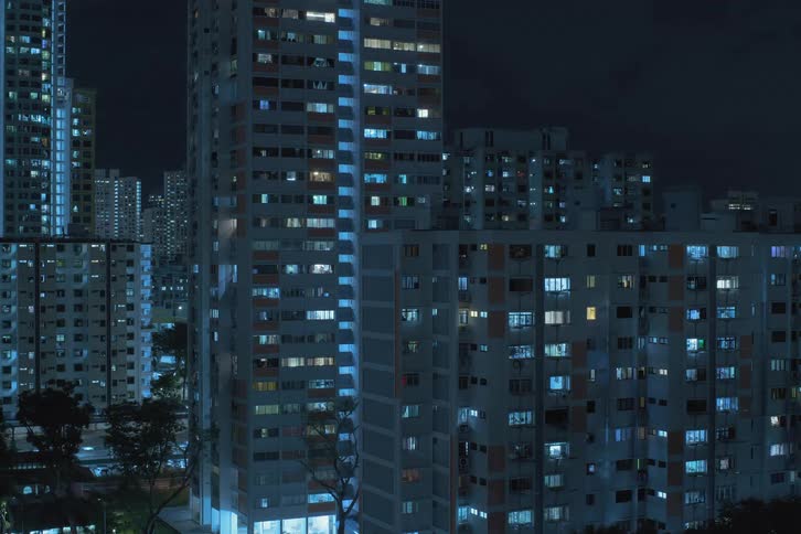A nighttime view of urban apartment buildings filled with people turning on window lights