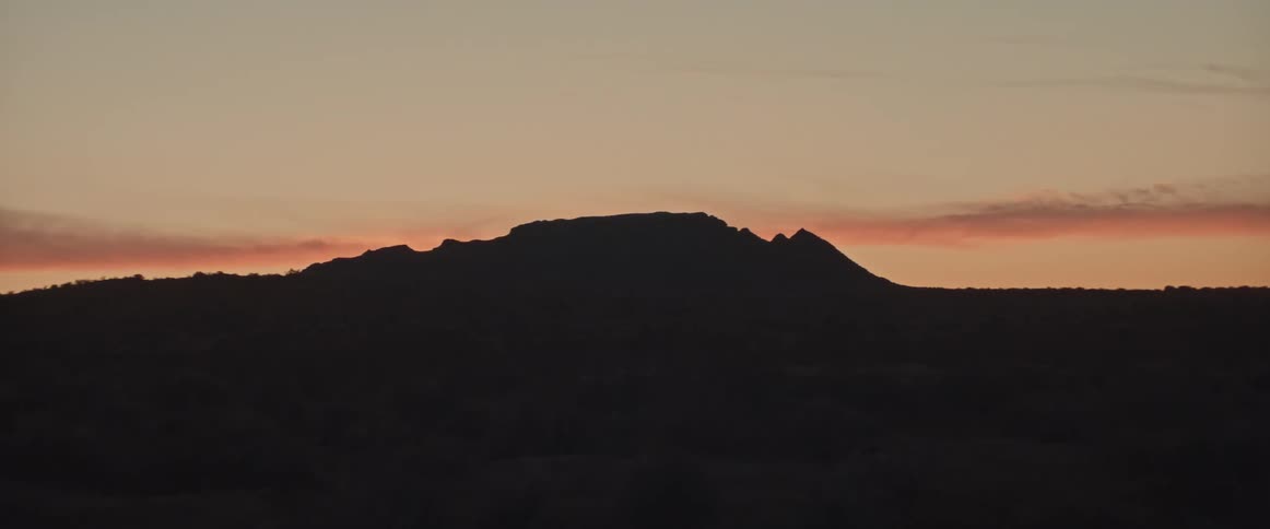 The camera zooms in on a mountain in the desert while the sun sets behind it