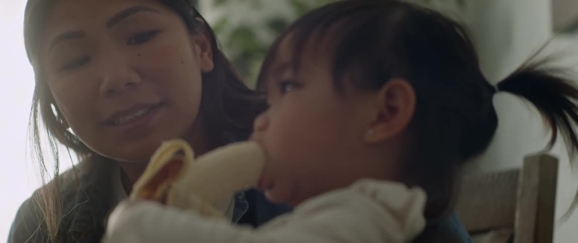 A little girl eating a bananas while her mother sits next to her at a table