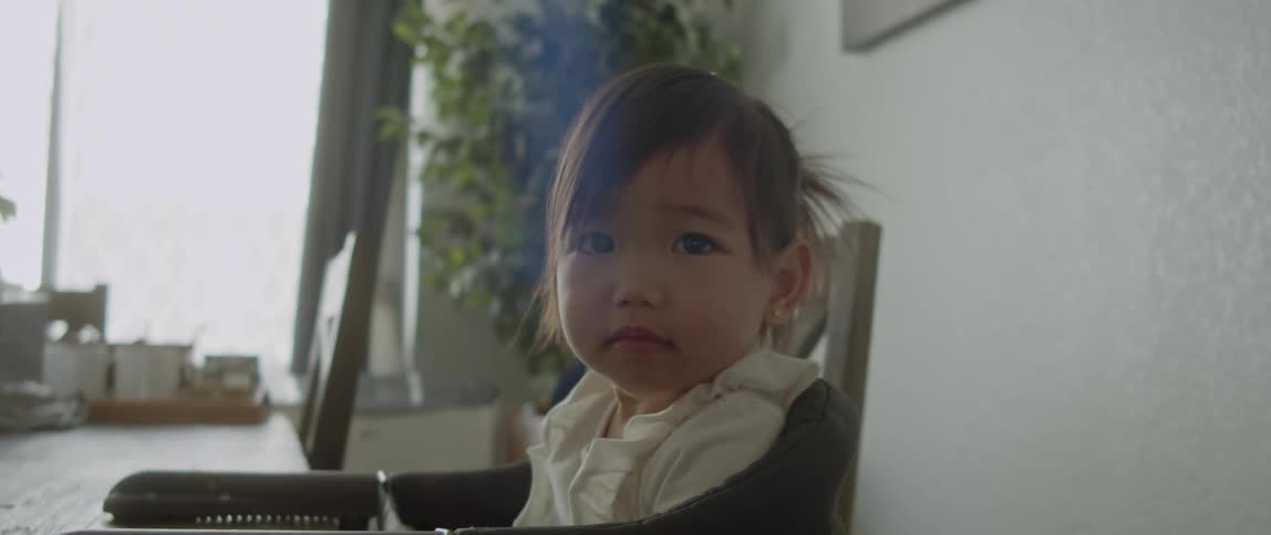 Toddler girl with white shirt and ponytail sits at table looking around