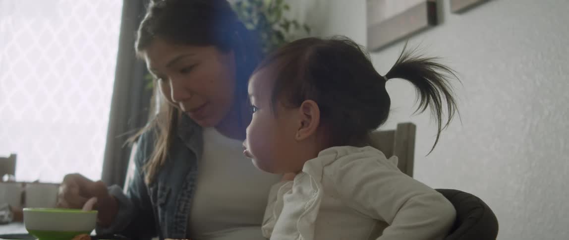 Mother with denim jacket and white top feeds toddler daughter sitting at table