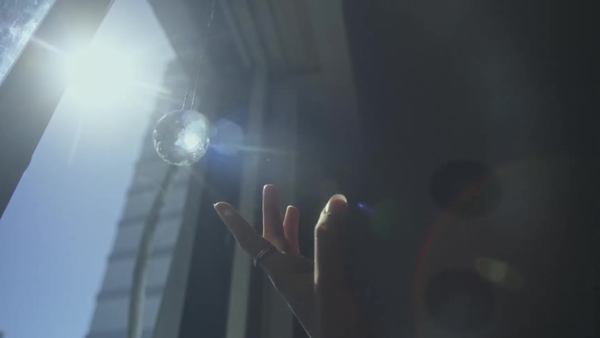 Close up of the extended fingers on the hand of a woman touching a hanging crystal ball hanging from a window in the sunlight