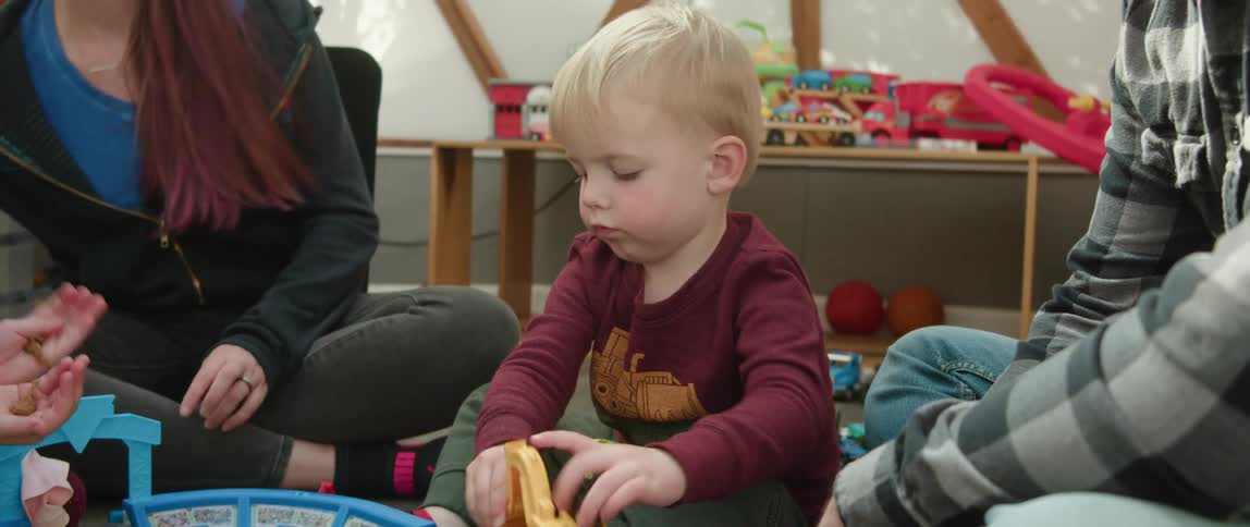 A toddler boy sitting with his mother and father plays with a plastic scoop loader truck on the floor