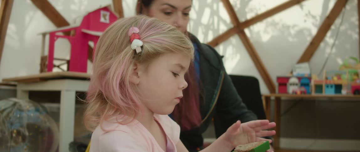 Girl with her hair tinted pink sit on the floor of a room playing with toys at daytime with her mother