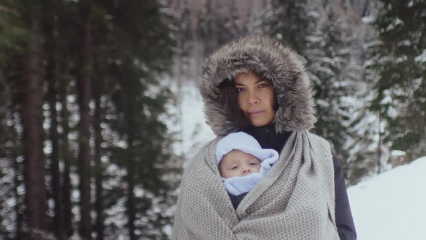 Mother stands looking to camera in the forest at winter with her newborn baby wrapped to her chest