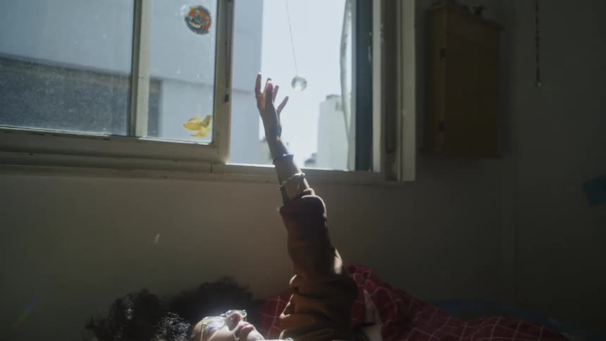 Young girl resting in bed touches a small crystal ball hanging from the window reflecting sunlight