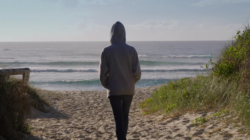 Woman with gray hoodie and black pants walks to the sea on the beach