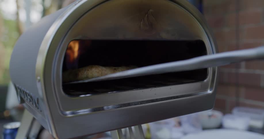 A hand uses a metal paddle to slide a pizza into an outdoor pizza oven at a cookout