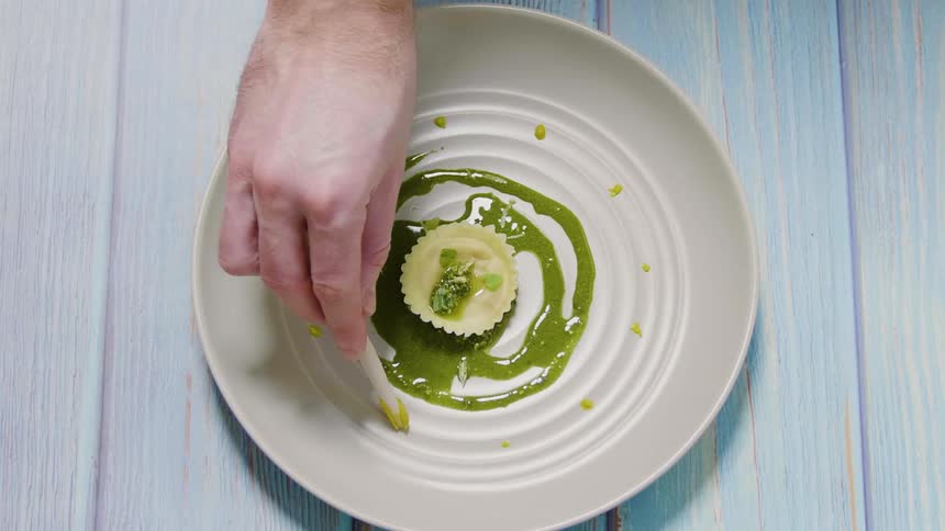 A mans hand pipes droplets of sauce around a single ravioli placed on top of green sauce on a plate