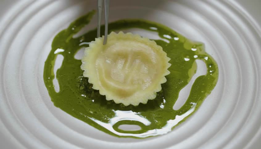 Someone uses tweezers to place a circular ravioli on top of a green sauce in a bowlshaped plate