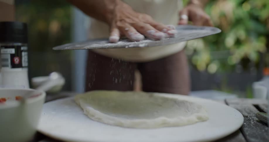 Close up of the hands of a man sprinkling flour over a pizza paddle on the backyard