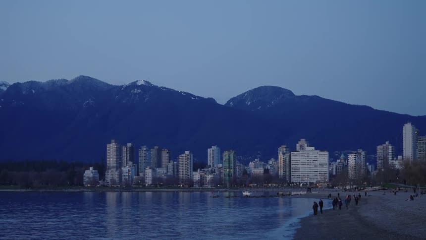 Landscape of people on the beach with high rise buildings and mountain range in the background at dusk