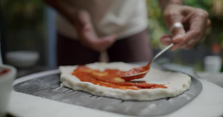 A person uses a spoon to spread sauce on top of dough on a table during the day