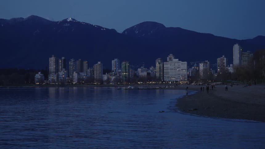 People walking on a beach next to a waterfront city in the mountains at evening