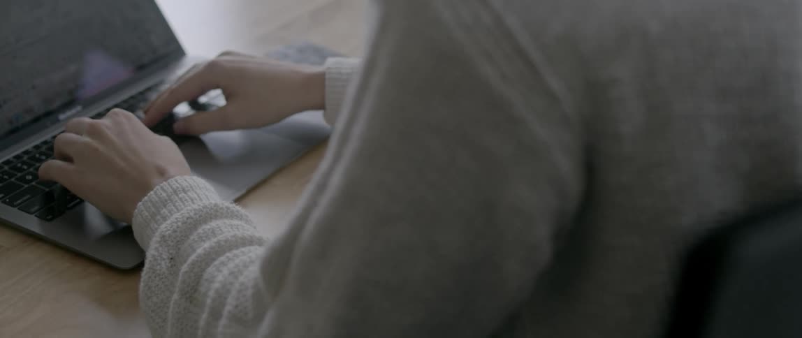 A woman in a sweater types on a laptop while sitting at a table