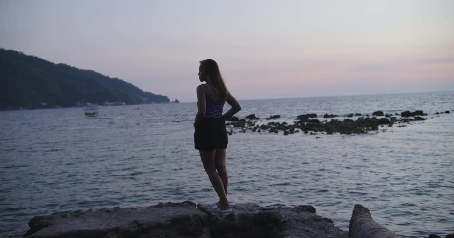 A woman stands on a rock overlooking the ocean at dusk